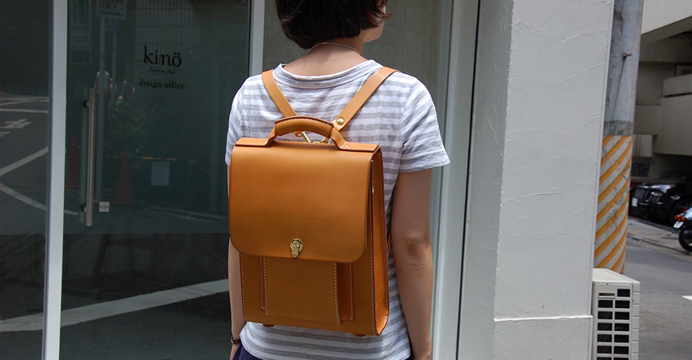 卡其色皮革復古後背包，具有頂部提把、翻蓋式前袋及金屬旋鎖，肩帶為皮革材質，與條紋上衣搭配。