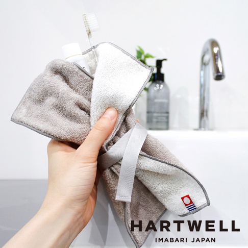 一條灰色毛巾，內側為白色，有灰色羅紋綁帶，一端露出牙刷和牙膏。毛巾印有 HARTWELL IMABARI JAPAN 標誌。