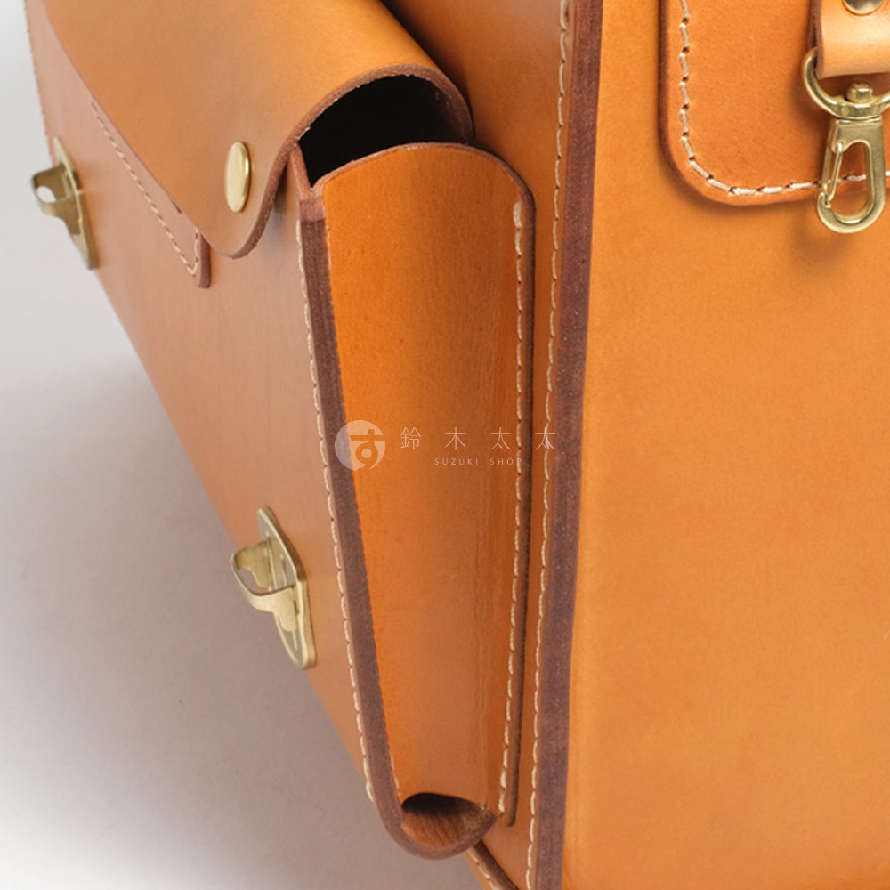 職人手作皮革橫式書包，採用天然皮革材質，呈現溫潤的焦糖色澤，搭配復古銅色金屬扣環與縫線細節，展現精緻工藝。