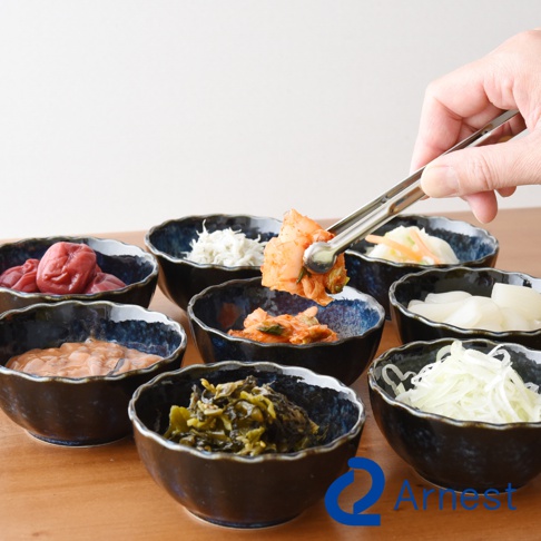 逸品物創不鏽鋼萬用料理夾，銀色，用於夾取泡菜，下方為裝有醃漬物（如醃漬過的梅子、小魚乾、泡菜、醃漬過的白蘿蔔、醃漬過的芥菜等）的深藍色波浪邊小碗。