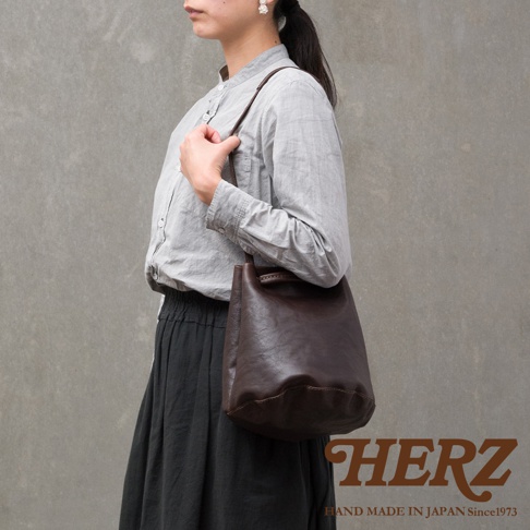 一位女性側面站立展示日本HERZ品牌深咖啡色皮革抽繩肩背托特包，該包款呈現自然柔軟的圓底桶狀結構，搭配細長皮革肩帶，整體風格簡約且具有手工質感，背景為單純的灰色牆面。