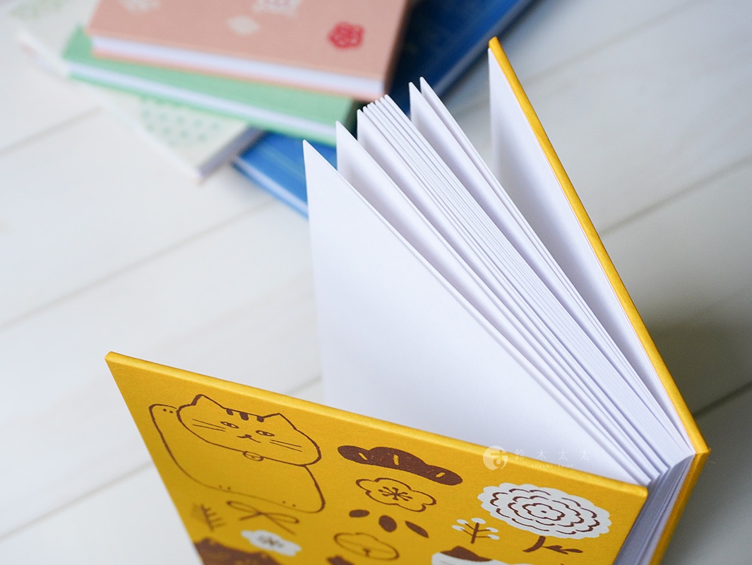一本鮮黃色封面的御朱印帳，封面印有可愛的招財貓與日式圖案，內頁為白色紙張，整齊排列。