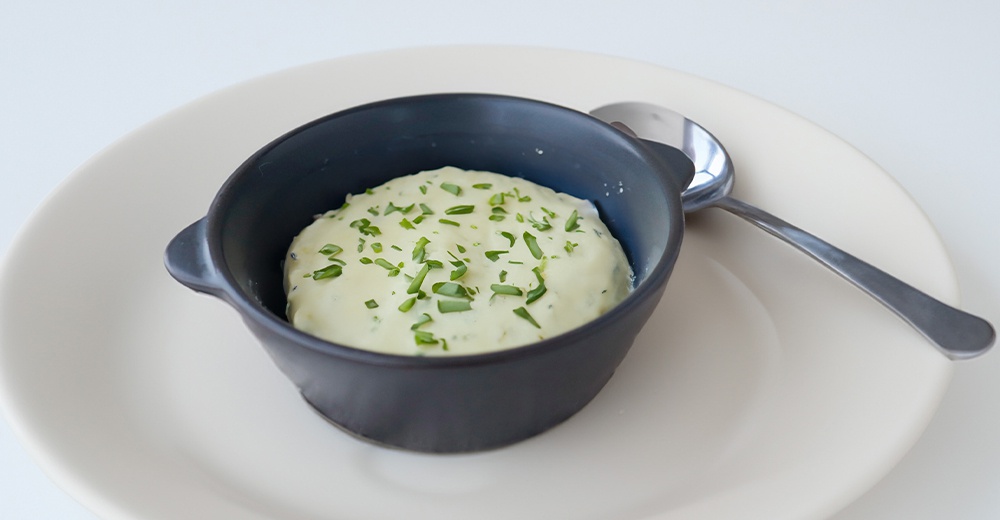 深灰色陶製迷你圓鍋，裝有撒上蔥花的奶油狀食物，放在淺色圓盤上，旁有湯匙。