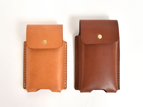兩個職人手作皮革手機收納包，分別為淺棕色和深棕色，均為翻蓋設計，配有金色按扣，側邊縫線清晰，底部有手機抽取開口。