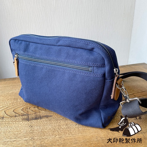 一個深藍色石洗帆布製成的旅行側背包，包身呈現方形，正面有兩個拉鍊口袋，其中一個較大，另一個較小，皮質的吊牌掛在拉鍊頭上，包包的背帶為黑色，旁邊有一個印有狗和包包圖案的商標，商標下方有中文文字。