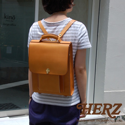 以橘棕色皮革製成的復古後背包，配有翻蓋和金色鎖扣，由一位女性背著，她穿著條紋上衣和深藍色褲子。