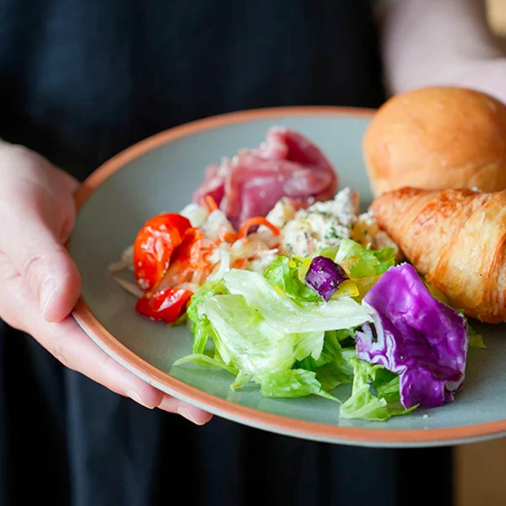 灰綠色陶瓷主菜盤，盤緣帶有橘色滾邊，盛裝著沙拉、番茄、肉片、麵包和可頌。