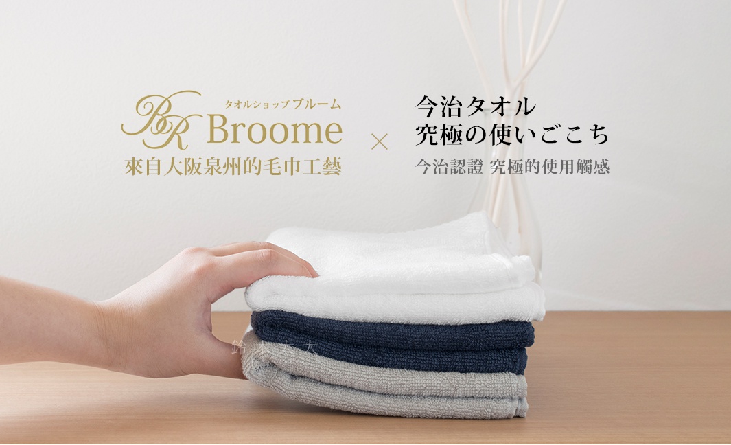 一手拿著一疊由白色、深藍色和灰色組成的今治純棉長毛巾，毛巾質感柔軟，表面有細微的菱紋。