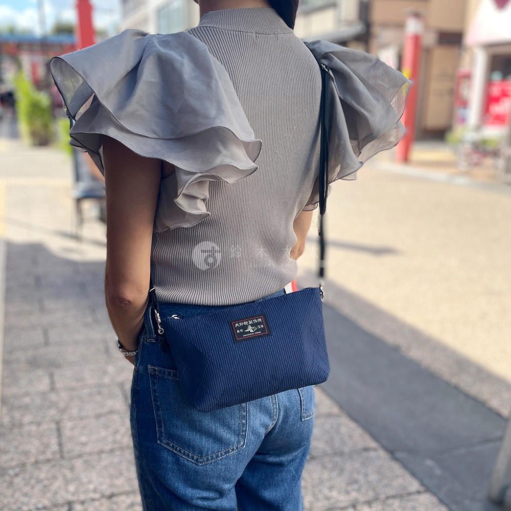 深藍色燈芯絨材質的隨身斜背包，斜挎於身著灰色荷葉袖上衣的模特兒身上，包包上有一個寫有日文的方形標籤。