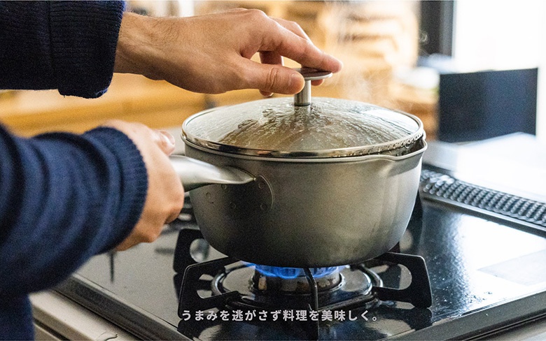 一個不鏽鋼雪平鍋，鍋身為銀灰色，配有透明玻璃鍋蓋和金屬手柄，正放置在瓦斯爐上加熱，鍋蓋上冒出蒸氣。