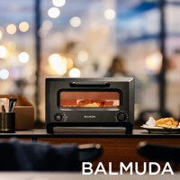 ReBaker 烤麵包機，採用黑色金屬外殼，設有透明玻璃門，內部有正在烘烤的食物。機身正面有黑色旋鈕和按鈕，以及 BALMUDA 品牌標誌。