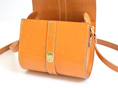 橘色皮革製成的單肩包，有著金屬皮扣和手工縫線，搭配皮革肩帶。