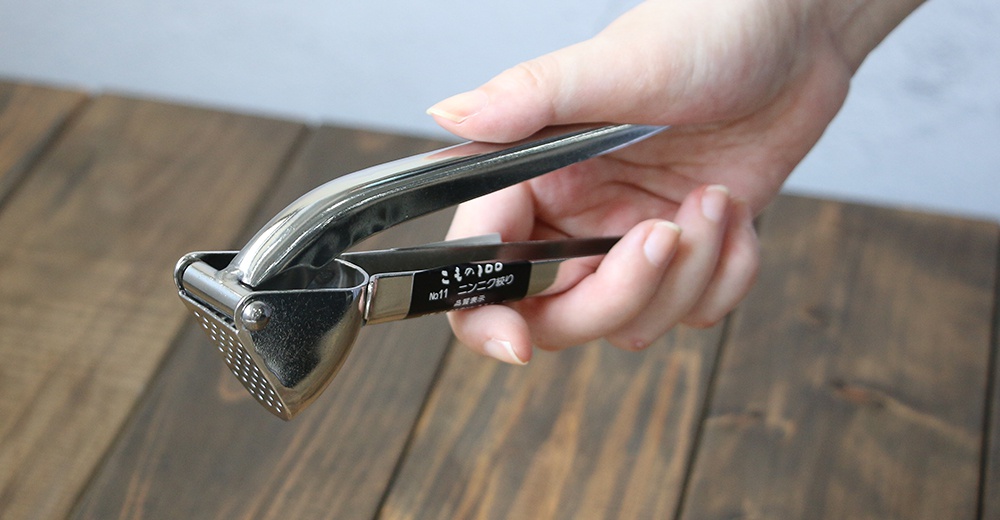 一只手正握著一個銀色不鏽鋼壓蒜器，壓蒜器底部有一個帶有許多小孔的蒜泥槽，以及兩個長柄。