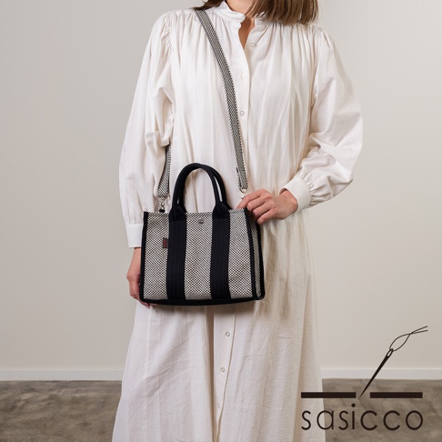 女性身穿白色洋裝，手提斜背一個黑白條紋的三河木綿刺子織方形托特包，包包有雙提把及可拆卸斜背帶。
