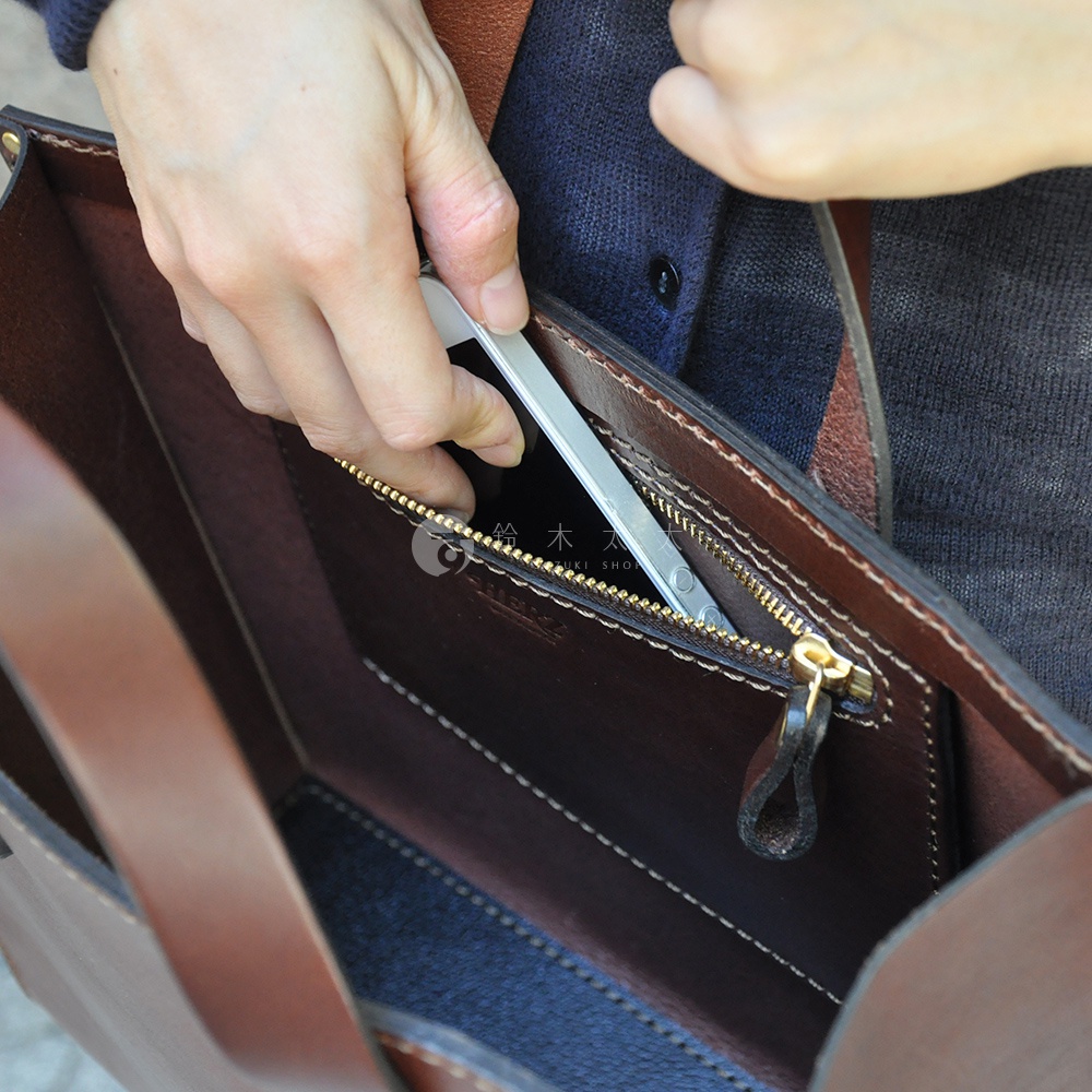 深棕色皮革直式托特包，內部有拉鍊暗袋，手機正被放入其中，包包帶有縫線細節。