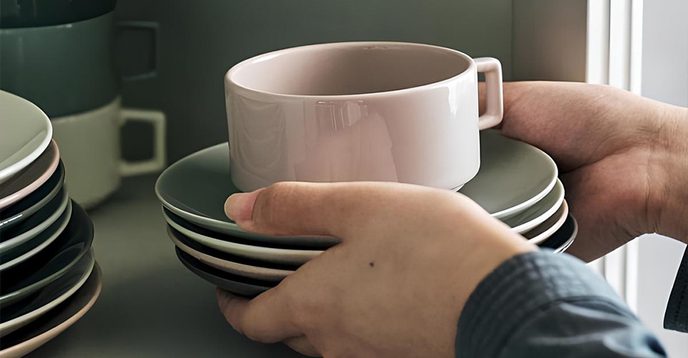 雙手捧著一疊深灰色的美濃燒圓形餐盤，最上方餐盤上放著一個粉色的圓形馬克杯。