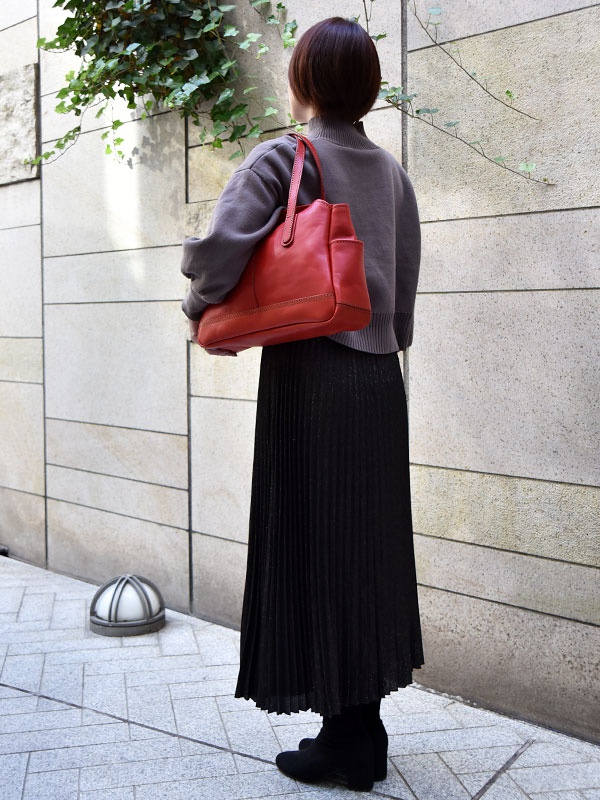 一位女性穿著灰色毛衣和黑色百褶長裙，肩上斜背著一個紅色皮革橫式托特包。包包有兩個側袋和一個頂部開口，質感光滑。