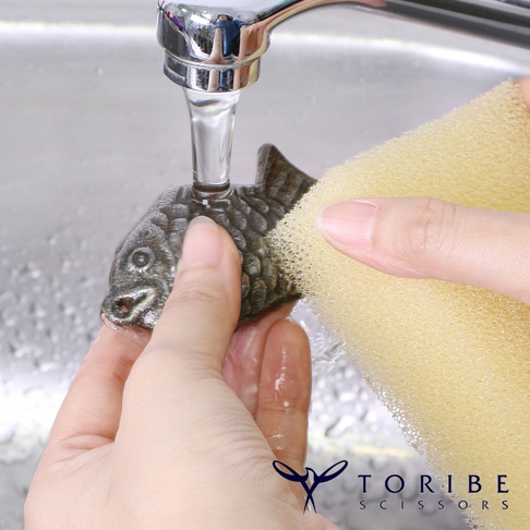 一隻深灰色、鑄鐵材質的鯛魚造型補鐵塊，在水龍頭下，由一隻手持著黃色海綿清洗。