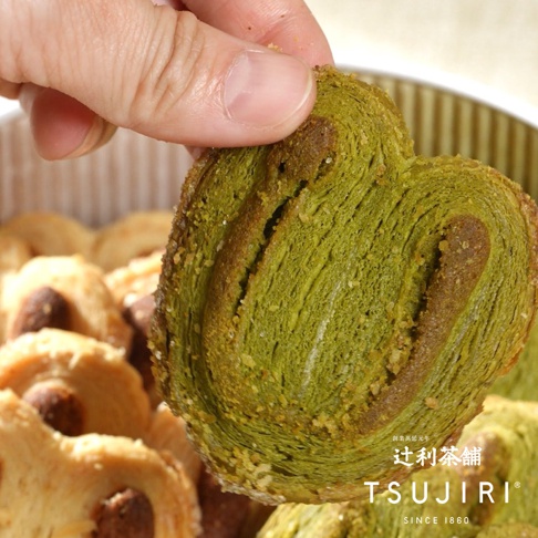 一隻手正捏著一片抹茶口味的蝴蝶酥，翠綠色的酥皮層次分明，邊緣帶有細緻的烘焙質感，背景為裝滿同款蝴蝶酥的鐵盒，角落處印有辻利茶舖的品牌標誌與創立年份文字。