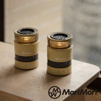 兩款淺黃色圓柱形無線藍牙音響，頂部有黑色網狀喇叭，中央有透明罩，罩內有 LED 燈發出暖黃光，外觀有水珠，桌面上有水漬。