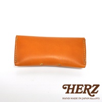 一個橘色的皮革眼鏡收納盒，上面有兩個黃銅色的按扣，底部有HERZ品牌標誌及日文手作字樣