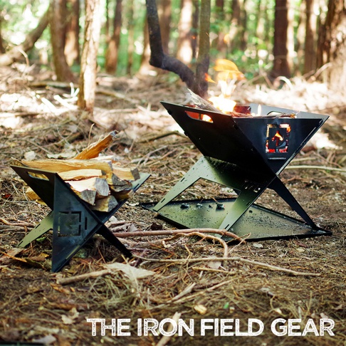 TAKI BEITA 焚火台爐架套裝 S，由黑色金屬製成，設計簡約，以「X」型支架支撐。其中一個焚火台正在燃燒火焰，另一個則堆放著木柴，兩者皆置於林間空地。