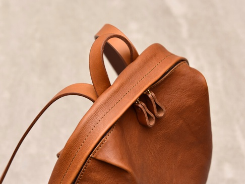 職人手作皮革後背包，採用溫暖的棕褐色，展現天然皮革的細緻紋理與簡約褶皺設計。雙拉鍊頭設計，搭配堅固的縫線，強調細節與質感。