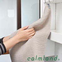 一雙手正在拿著一條米色無撚紗質地的長方巾，其表面呈現細緻的織紋。毛巾掛在金屬掛鉤上，背景為白色櫃子和門。