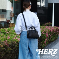 一位女性顧客在盛開粉紅色杜鵑花叢旁，身穿寬鬆白色連帽衫和牛仔褲，斜挎一個黑色皮革手提肩背包，包包上有HERZ品牌標誌，背景為建築物。