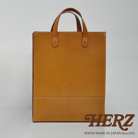 職人手作皮革直式托特包 T-44 L，採用堅韌的天然皮革製作，呈現自然的焦糖棕色。包款設計簡潔，配有雙提把，正面下方有兩道橫向縫線裝飾，整體風格經典實用。