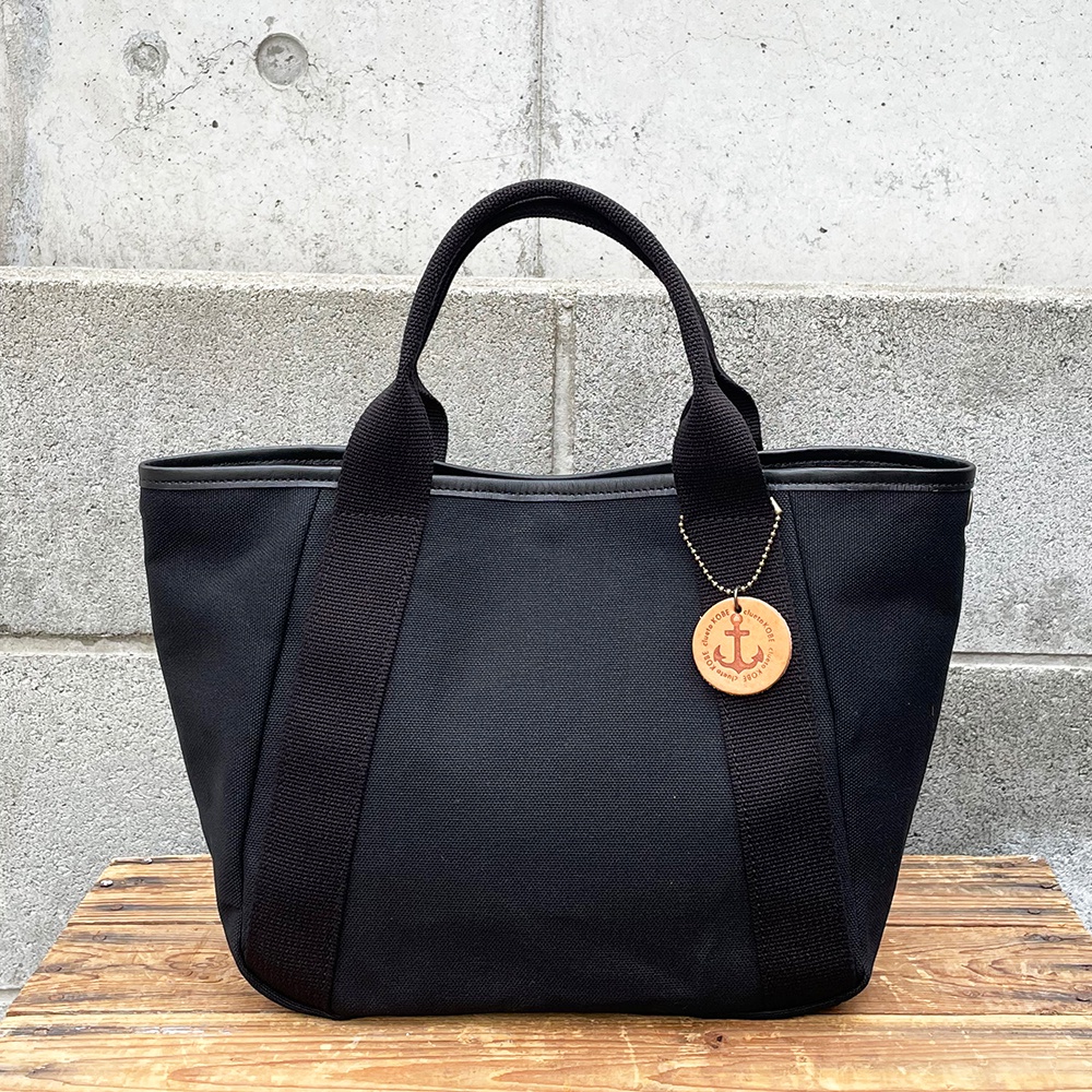 黑色帆布多口袋手提托特包，配有黑色織帶提把和皮革飾邊。包包上掛有圓形木質吊牌，上面印有船錨圖案和文字。