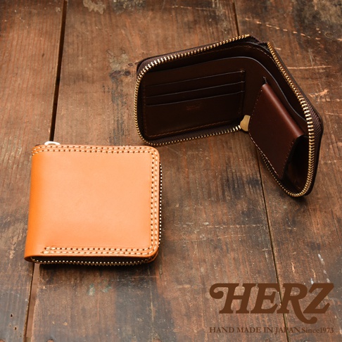 這張照片展示了日本HERZ職人手作皮革短夾，左側為一個淺褐色皮革短夾，外圍縫線細緻，右側則為另一個已打開的深褐色短夾，呈現出內部多層卡片收納槽與硬幣袋的設計，整體擺放在木質桌面上，右下方印有HERZ品牌字樣與產地資訊。