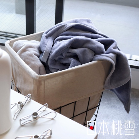 一條淺藍色、柔軟的舒眠雙面四季毯，疊放在一個米色帆布內襯的儲物籃中，旁邊還有另一條淺色毯子。