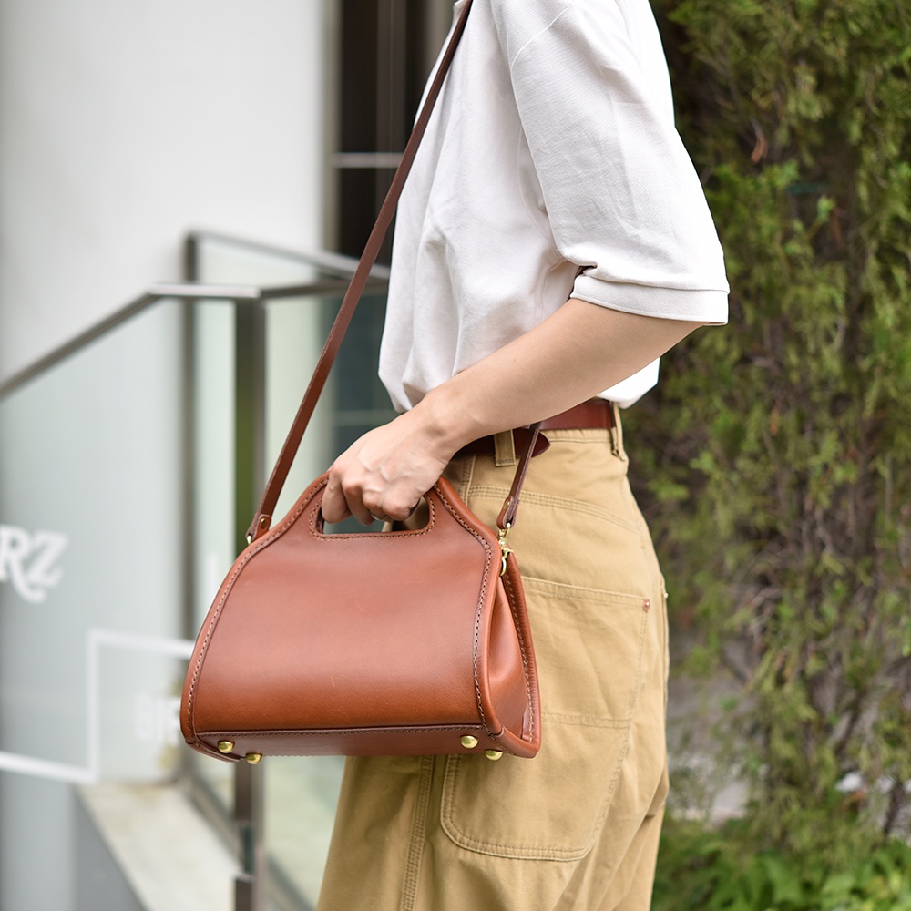 職人手作皮革單肩兩用拱型手袋，採用棕色皮革製成，袋身呈現弧形，配有提把和可拆卸的長肩帶，底部有金屬腳釘。