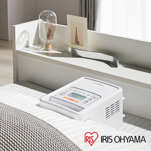 白色IRIS OHYAMA強力被褥乾燥機 FK-H1，配備液晶顯示螢幕和多個操作按鈕，位於鋪有灰白色條紋床單和灰色針織蓋毯的床鋪上。