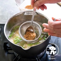 不鏽鋼雪平鍋內煮著綠色蔬菜和湯，湯面上飄著少量泡沫。一隻手正用湯匙將味噌醬透過濾網加入鍋中。