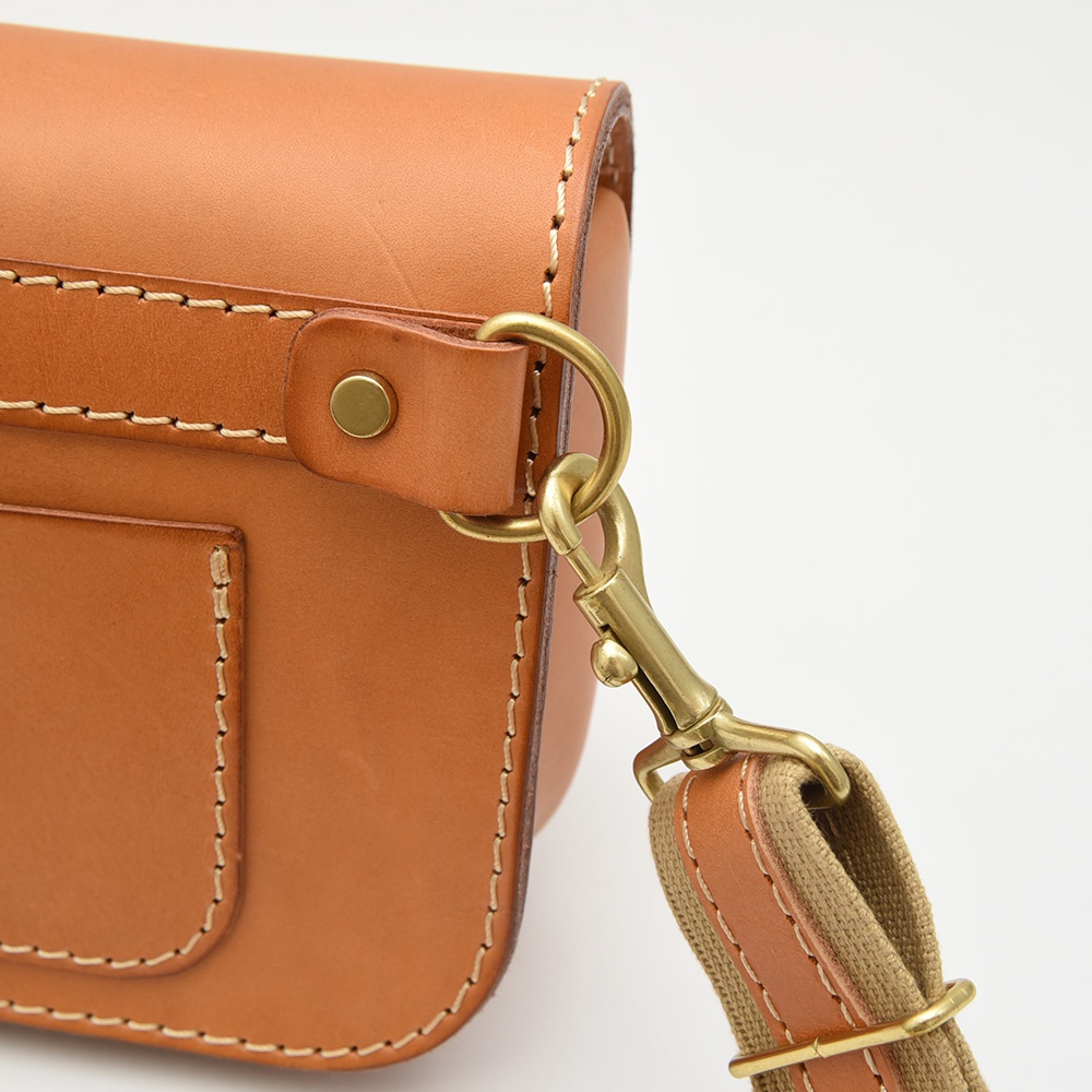 職人手作皮革單扣經典斜背包，採用淺棕色皮革，搭配黃銅色金屬扣環與旋轉鉤，可調節織帶與皮革拼接的斜背包帶。