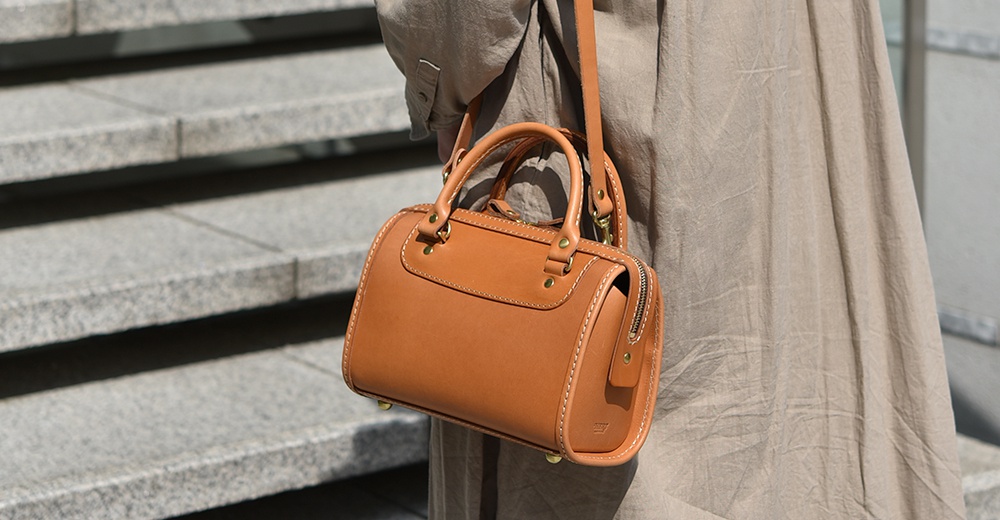 職人手作皮革迷你波士頓包，呈現溫潤的焦糖棕色，採用光滑皮革材質，搭配金色金屬拉鍊和鉚釘，設計簡潔，附有提把與可拆卸斜背帶。