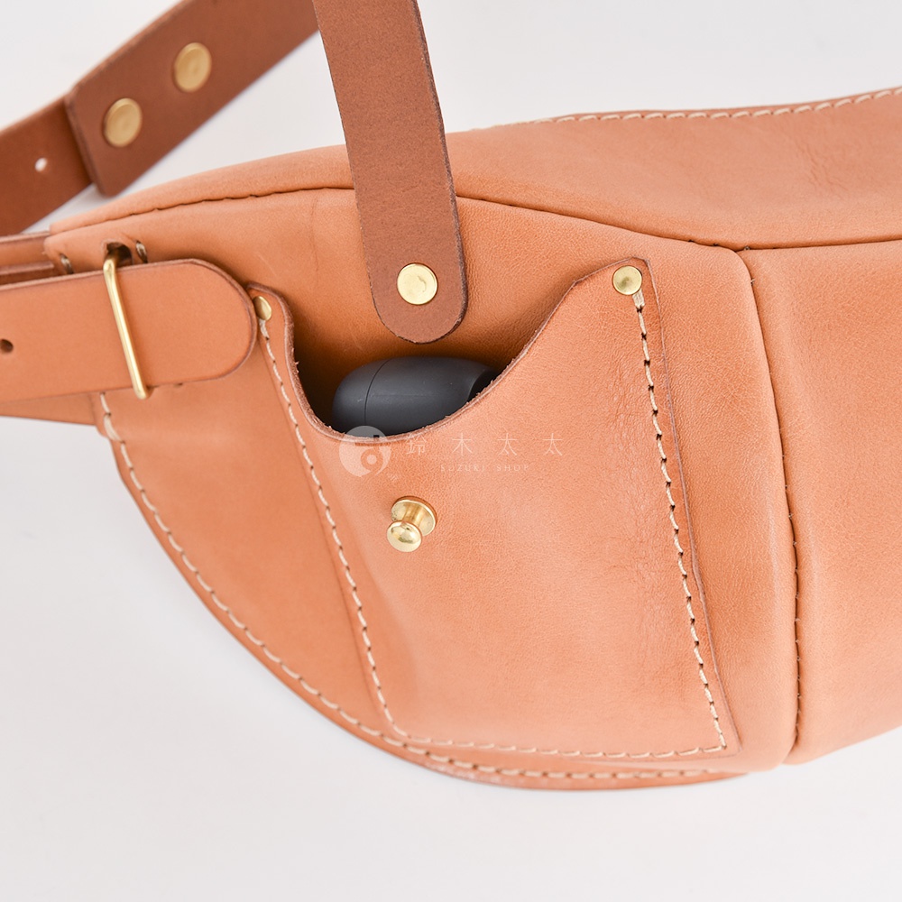 日本HERZ職人手作皮革大容量斜背包F-1，特寫展示包款一角，皮革呈現自然光澤，包身設有方便收納小物的小口袋，口袋內可見一組黑色的無線耳機，口袋外觀有黃銅色旋鈕扣件。