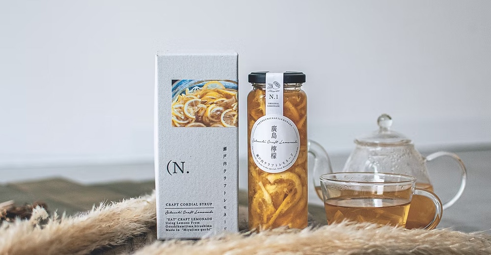 這張圖片展示了一瓶廣島手工熟成檸檬釀與其包裝盒，右側放有一杯沖泡好的檸檬茶與玻璃壺，前方以乾草裝飾，整體呈現清新且充滿質感的日式氛圍。包裝盒與瓶身上印有 N.1 與瀨戶內手工檸檬釀等文字，強調使用嚴選檸檬與職人手作特色。