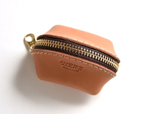 一個棕色皮革製貝殼造型零錢包，配有金色拉鍊和皮革拉環，表面有「HERZ」壓印。
