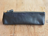 黑色皮革製成的職人手作 Organ 三角收納筆袋，配有金色拉鍊和一個皮革拉環，側面有品牌標誌，置於木紋桌面上。