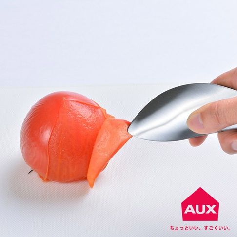 手持不鏽鋼調理夾，正在夾取一個正在被剝皮的紅色番茄。調理夾呈彎月形，表面光滑。番茄表皮被剝下一塊，露出橘紅色的果肉。背景為白色。