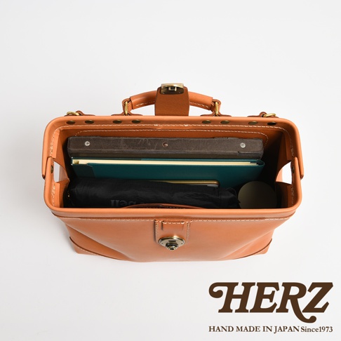 從上方俯視的角度拍攝，展示一只日本HERZ品牌職人手作的棕色皮革公事包。包身呈現硬挺的杜勒斯包結構，內部整齊擺放著記事本、黑色收納袋與文具用品。畫面右下方印有品牌名稱與手工製作年份標語。