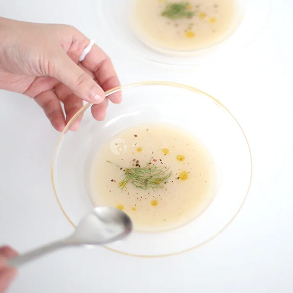 透明玻璃深型點心碗，內盛淺黃色湯品，頂部點綴香草和胡椒粒。一只手戴著白色戒指，正在拿取湯碗。