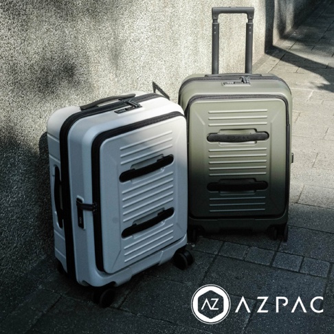 兩個 AZPAC 品牌的前開口設計行李箱並排置於灰色石板地面與牆面前，左側為白色款式，右側為帶有行李拖桿的綠色款式，兩者皆擁有橫條紋路外殼與黑色手把細節，整體呈現極簡俐落的工業風格。