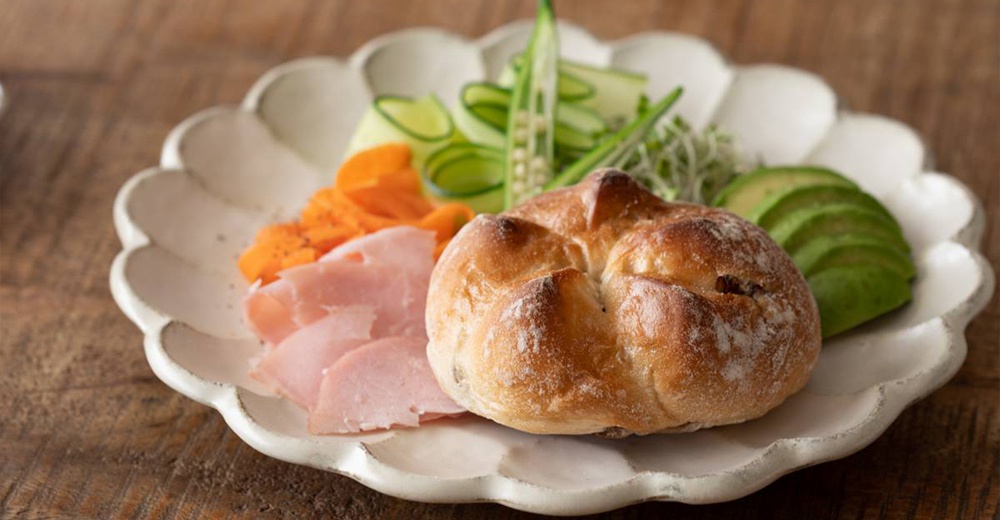 一個白色、帶有花瓣邊緣的陶瓷主食盤，盤中盛有圓形麵包、切片火腿、紅蘿蔔絲、黃瓜片、秋葵和酪梨片。