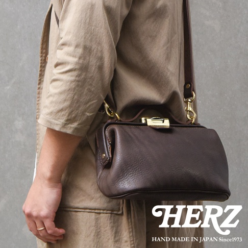 模式B： 揹著日本HERZ職人手作皮革Organ鎖孔式大開口肩背包M-7，包款為深棕色，材質為皮革，以側邊視角拍攝，包包以金屬鎖孔式開合，並有HERZ品牌標誌。
