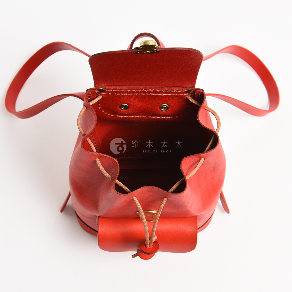 一款由日本HERZ職人手作的紅色皮革經典迷你後背包，採用堅韌的Stare皮革製成，頂部設有黃銅扣環與抽繩束口設計，包身呈現溫潤質感，上方印有鈴木太太標誌，整體造型簡約精緻。
