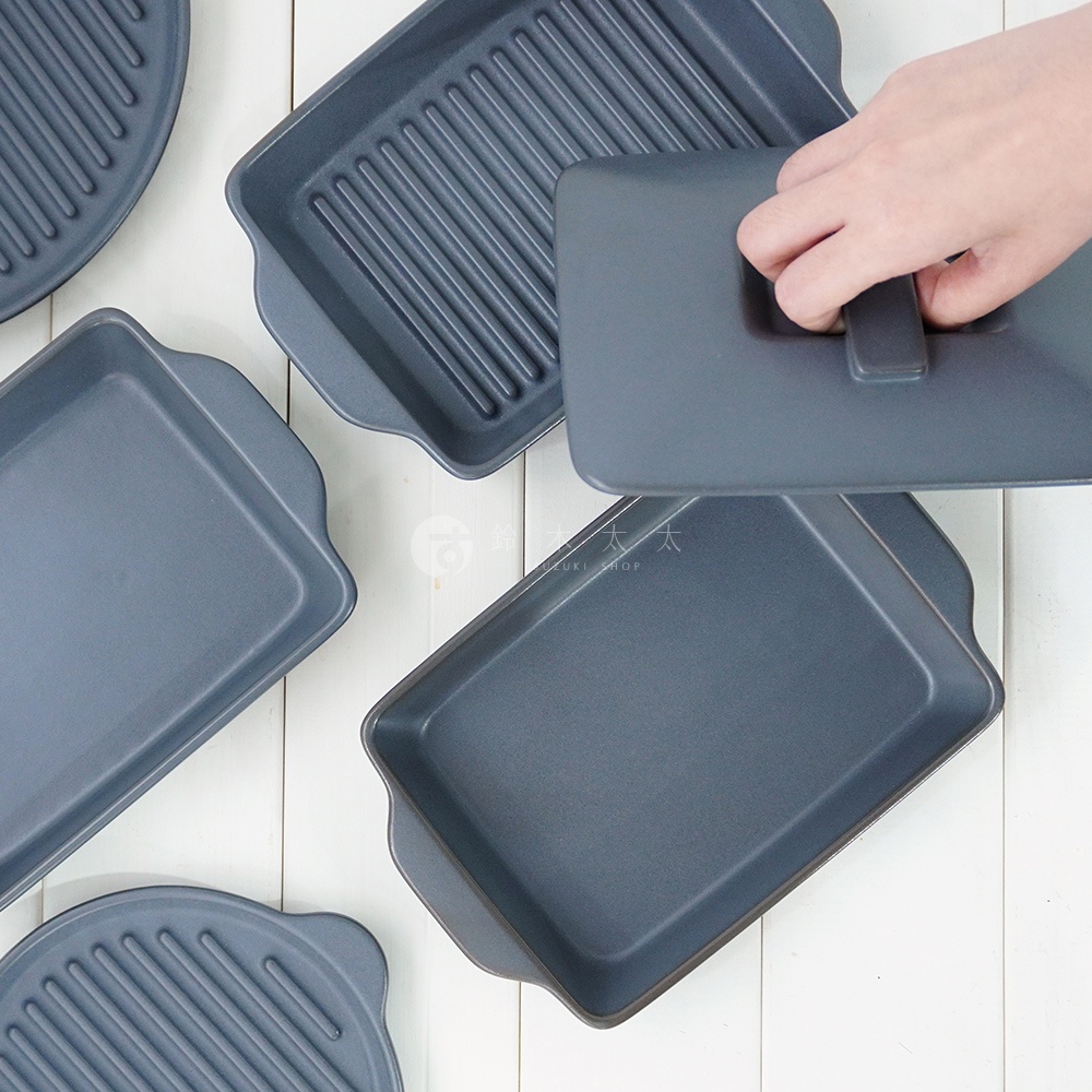 多個深灰色陶製烤盤，其中一個為長方形平面，另有兩個為長方形烤盤，其中一個有烤紋，一個蓋子被手拿起，整體呈平鋪狀。
